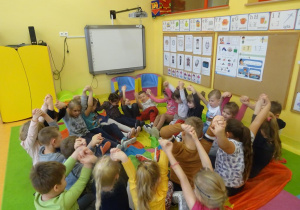 Grupa dzieci trzymająca się za uniesione dłonie siedzi w okręgu na chuście animacyjnej.